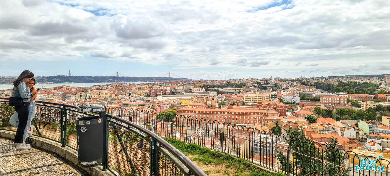 A scenic overlook with two people standing by a railing, offering a panoramic view of a city with a mix of historical and modern buildings, and a distant bridge spanning a river under a partly cloudy sky.