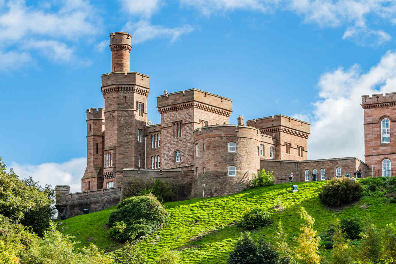 A historic castle with towering turrets and brick walls is situated on a grassy hill with trees and shrubs, under a bright blue sky with scattered clouds.
