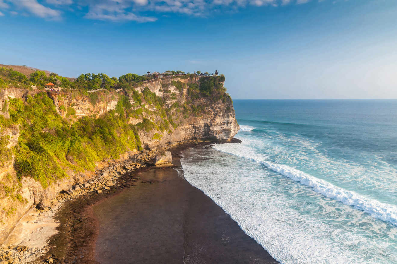 Coastal cliffside view with waves crashing against the rocky shoreline under a clear blue sky. Vegetation covers the top of the cliff.