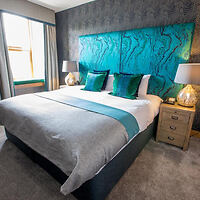 A hotel bedroom with a large bed featuring a teal headboard, grey and teal bedding, bedside tables with lamps, and a window with curtains.