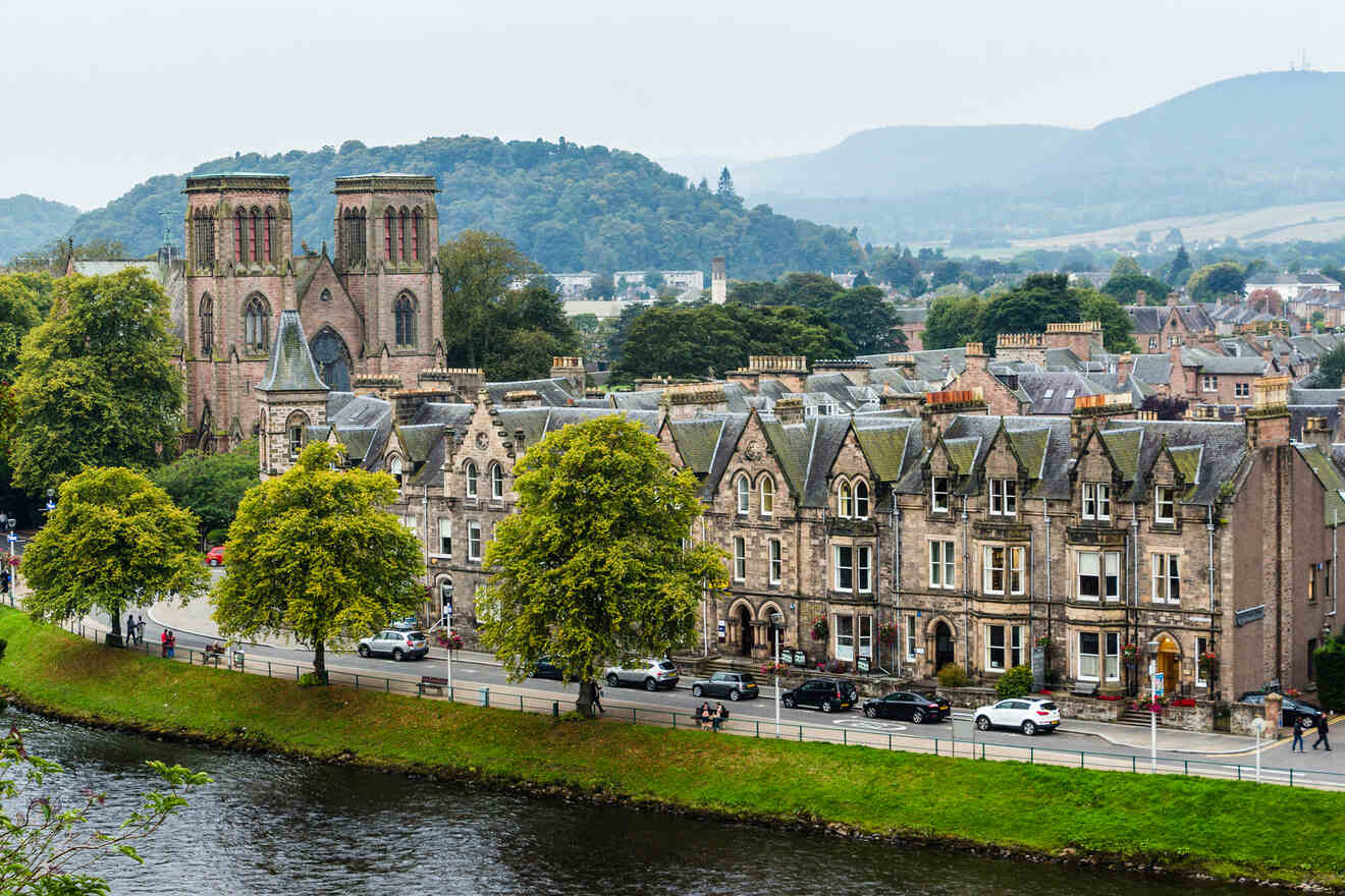 A row of historic buildings and a cathedral stand along a riverbank with lush green trees, with a backdrop of hilly landscape under an overcast sky.