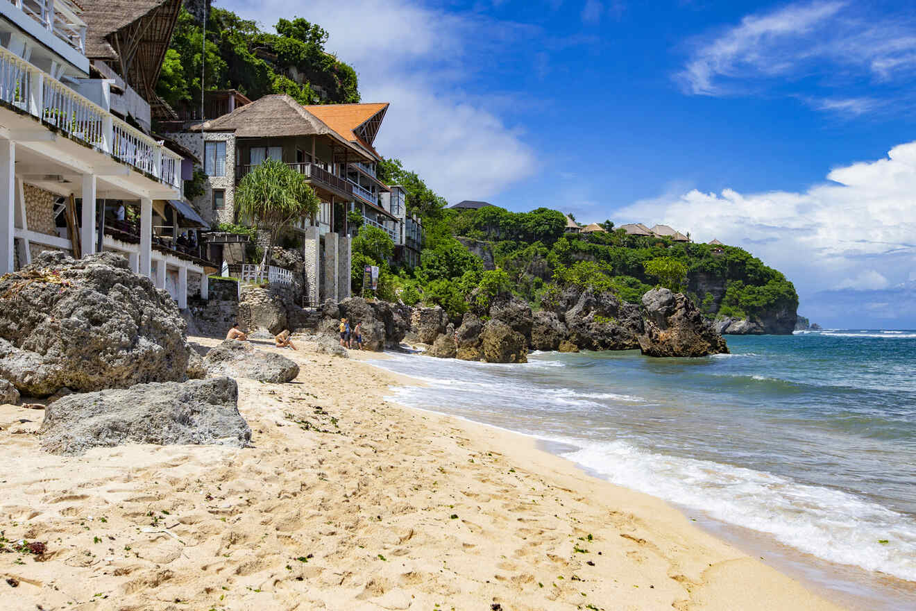 Sandy beach with rocky shoreline and ocean waves. Cliffside houses and lush greenery under a bright blue sky.