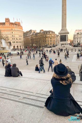 the author of the post in a black hat sits on steps overlooking a bustling public square filled with people, and buildings in the background. A tall column structure is also visible.