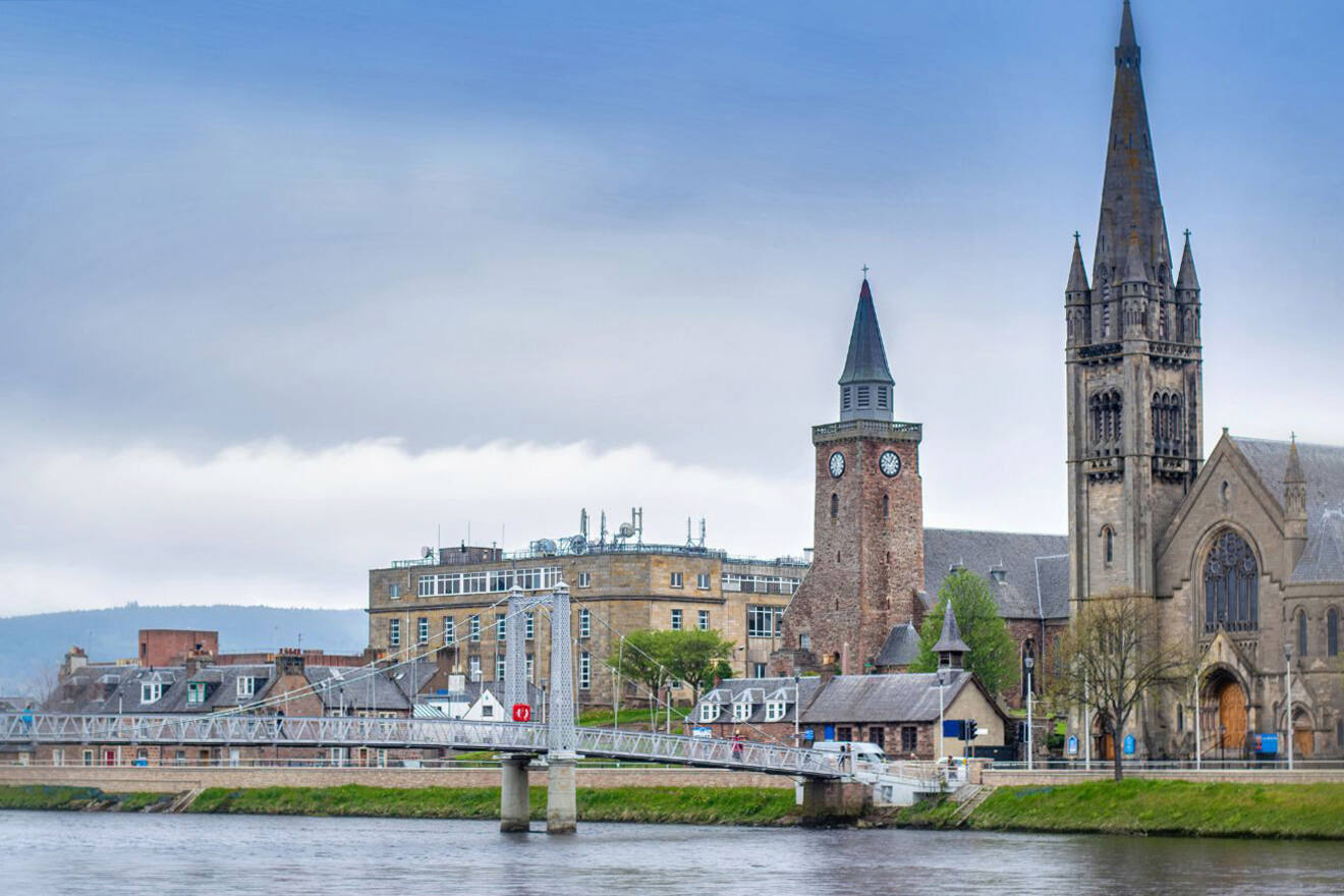 View of Inverness, Scotland, featuring a riverside scene with historic buildings including clock and church towers, seen under a cloudy sky with a pedestrian bridge over the River Ness in the foreground.