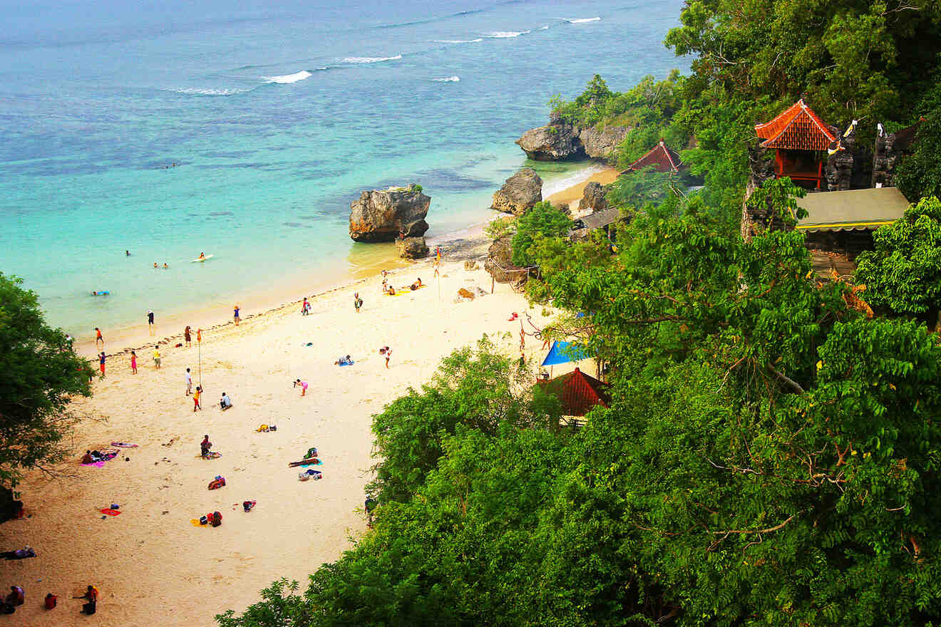 Aerial view of a beach with people, turquoise water, rocks, greenery, and a small building with a red roof.