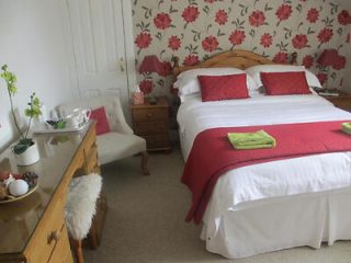 A neatly arranged bedroom with a double bed adorned with red pillows, a matching throw, and two green towels. The room features floral wallpaper, a wooden dresser, a chair, and nightstands with red lamps.