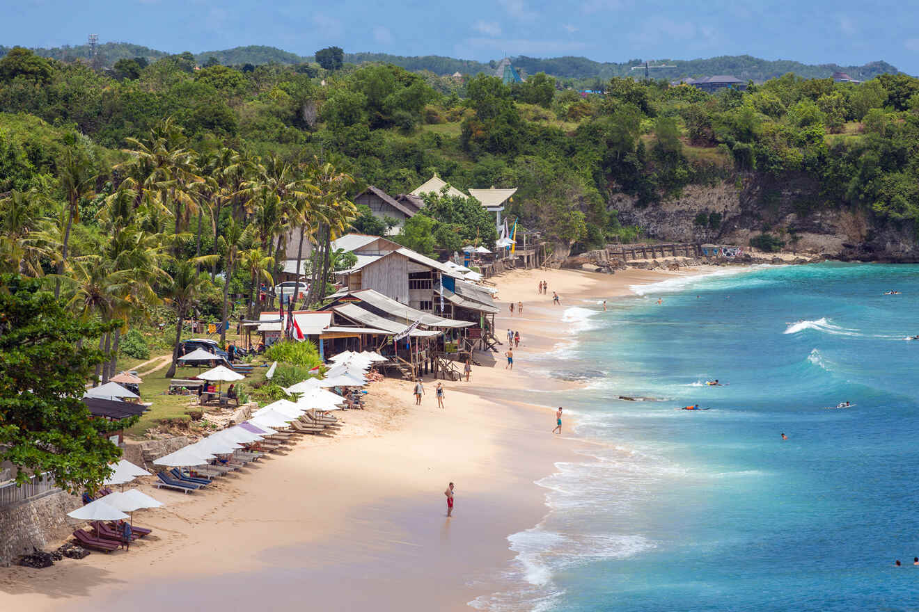 A sandy beach with people swimming and walking. Palm trees and buildings line the shore. White umbrellas and lounge chairs are set up along the beach. The ocean is a bright blue.