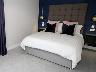 A neatly made bed with a white duvet, pink and black pillows against a blue accent wall, flanked by two hanging gold lamps and small bedside tables.