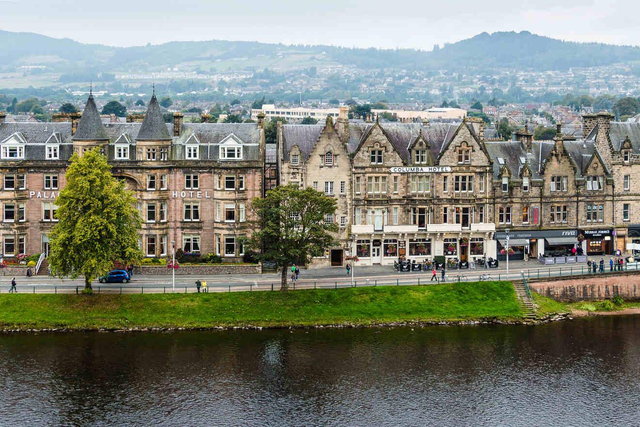 A row of historic buildings, including two hotels, lines a riverside in a town with hills in the background.