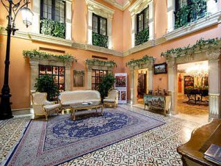 Elegant hotel lobby with patterned floor tiles, classic furniture, a large rug, and potted plants. Walls are peach-colored, and the space has a high ceiling with tall windows adorned with greenery.