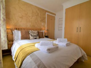A neatly arranged bedroom features a double bed with white and yellow linens, folded towels on top, and wooden wardrobes. A deer-themed cushion and two bedside lamps add decorative elements.