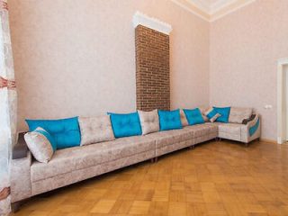 A long beige sectional sofa with blue cushions is arranged along two walls of a room with a brick column and parquet flooring.