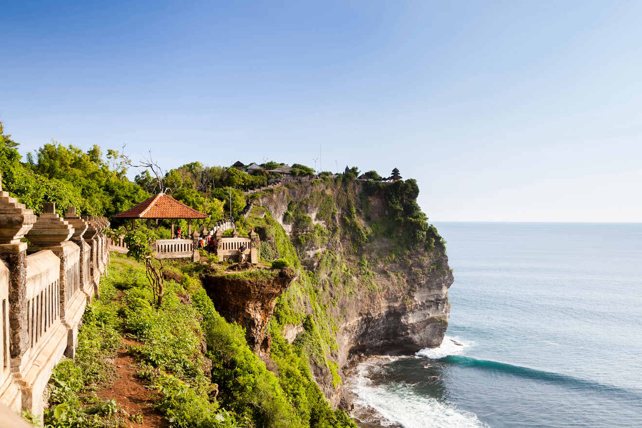 Cliffside view of Uluwatu Temple in Bali, Indonesia, overlooking the ocean, with lush greenery and a tiled-roof pavilion.