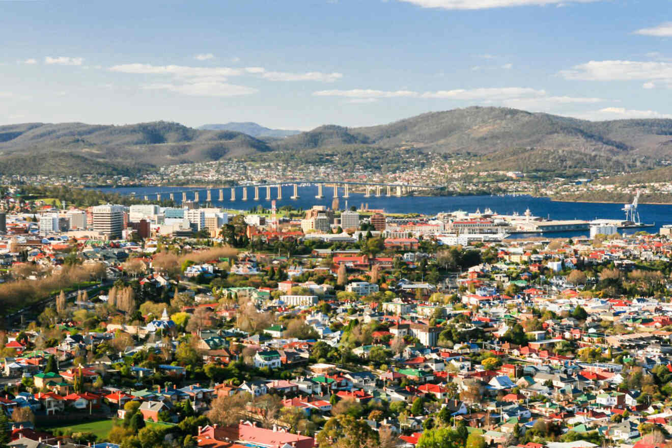 Aerial view of a city with colorful rooftops, a river, a bridge, and hilly landscape in the background under a partly cloudy sky.