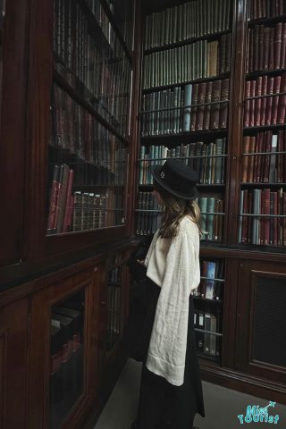 the author of the post wearing a black hat and a white sweater stands in front of a large wooden bookshelf filled with books, gazing at the volumes behind glass doors.