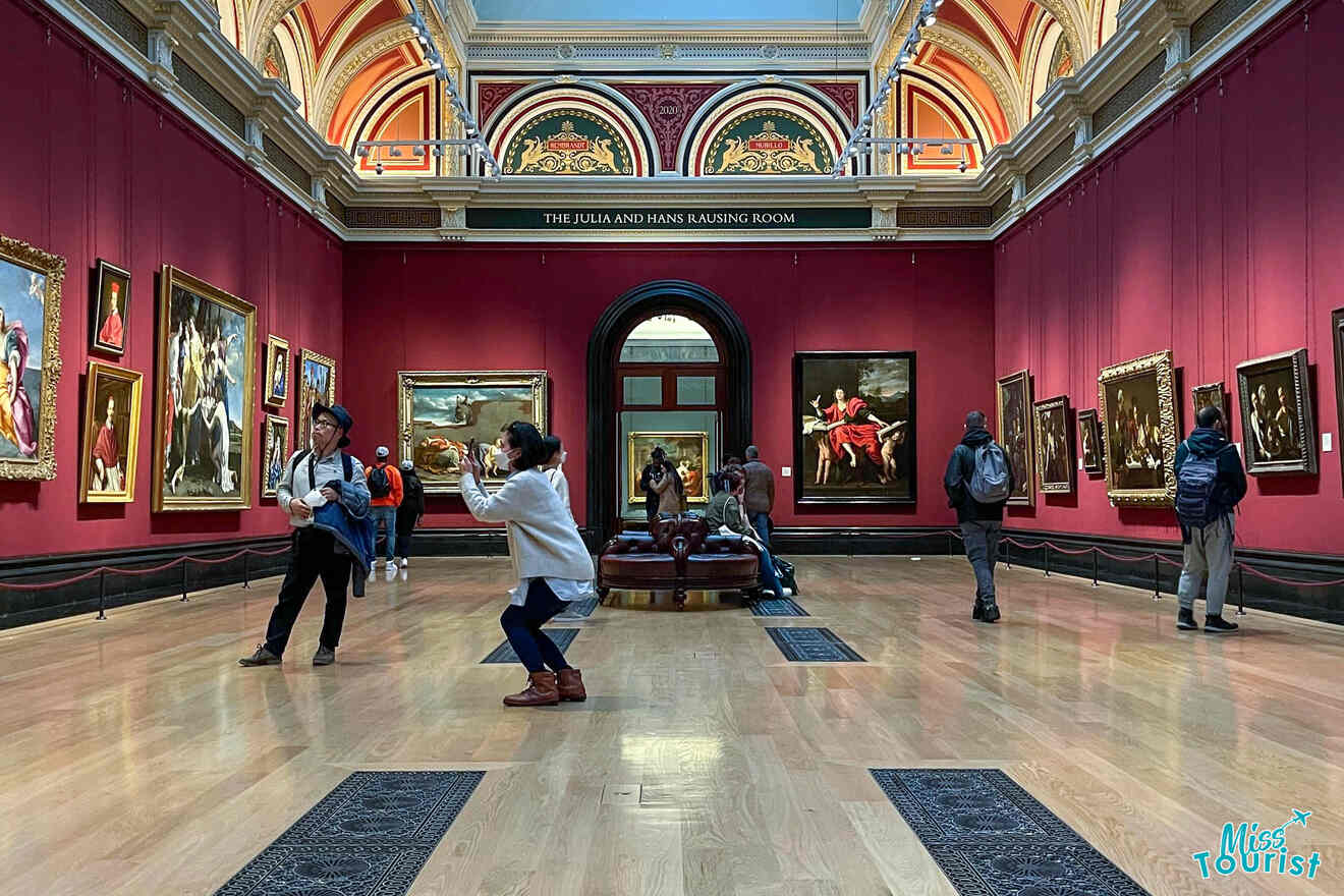 People are observing and photographing artwork in a gallery with red walls and ornate archways.