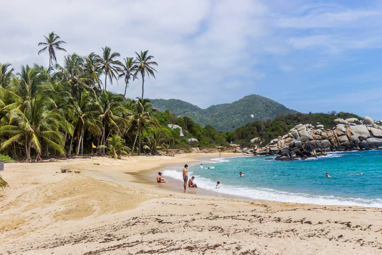 A sandy beach with people swimming and relaxing, lined with palm trees and surrounded by rocky outcrops and lush green hills under a partly cloudy sky.