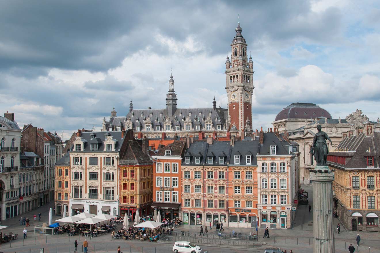 A historic European city square with colorful buildings, outdoor cafes, and a prominent clock tower under a cloudy sky.