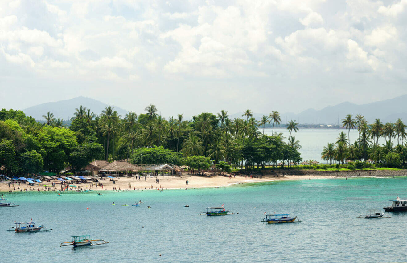 A tropical beach with palm trees, clear turquoise water, and several small boats. People gather on the sandy shore under a partly cloudy sky. Hills are visible in the background.