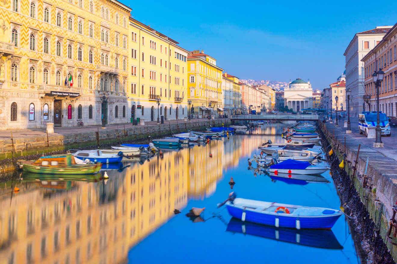 Calm evening view of the Grand Canal in Trieste, Italy, with colorful boats moored along the reflective water and classical buildings lining the sides