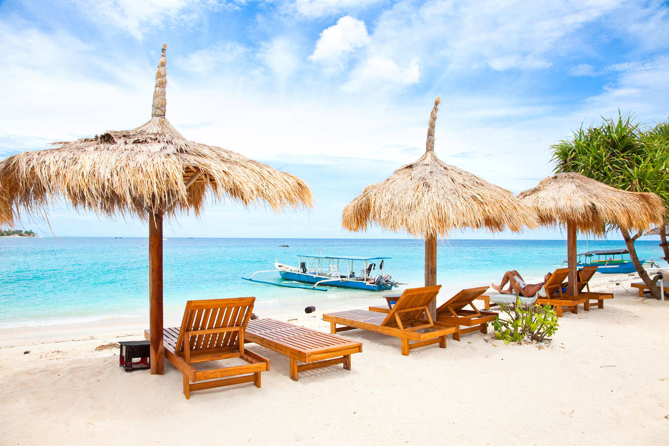 Beach scene with wooden lounge chairs and straw umbrellas on white sand, overlooking a turquoise sea. A boat is anchored nearby, and a person is relaxing on one of the chairs under clear skies.