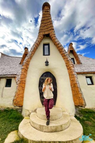 author of the post standing on stone steps in front of a fairy-tale style house with pointed roofs, wearing a beige sweater and maroon pants. Cloudy sky in the background.