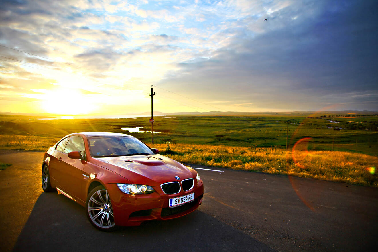 A red BMW sports car parked on a road, with a scenic background of grassy fields and a sunset sky.