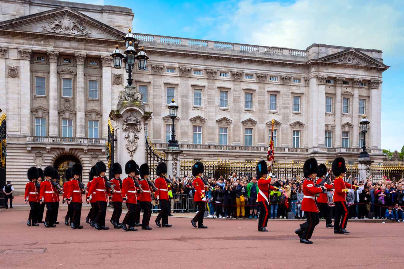 A group of guards in red uniforms and black hats march in front of an ornate building with a large crowd watching.