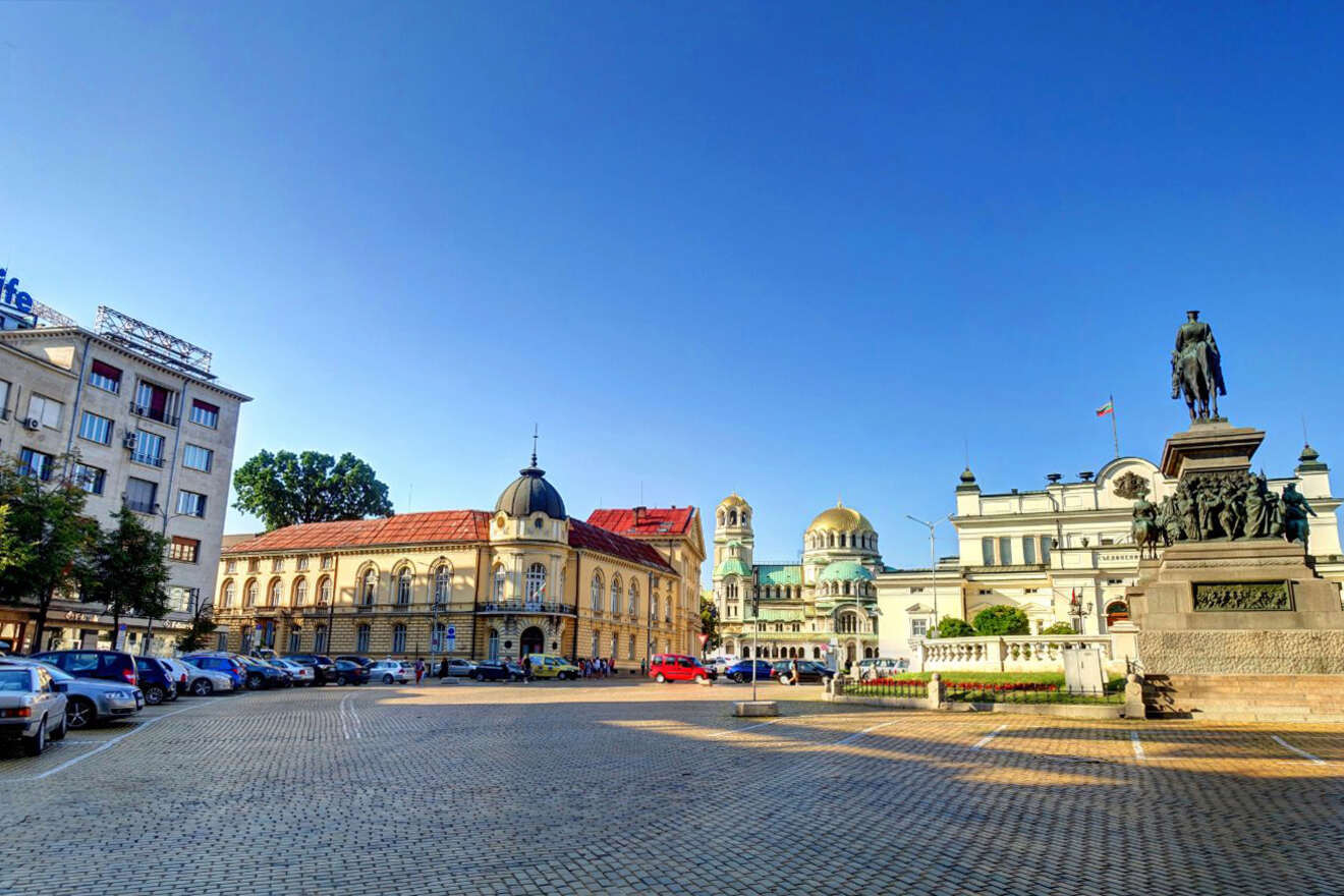 Urban square with parked cars, a statue, buildings with red and green domes, and a clear blue sky.