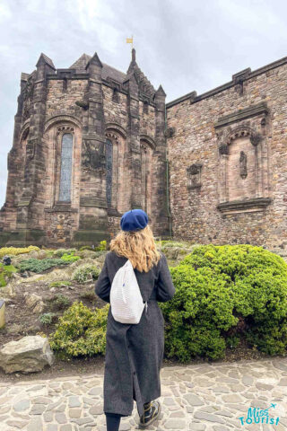 author of the post wearing a long coat and a blue beret stands with their back to the camera, facing a historic stone building. They carry a white backpack. Shrubs and stone paths are visible in the foreground.