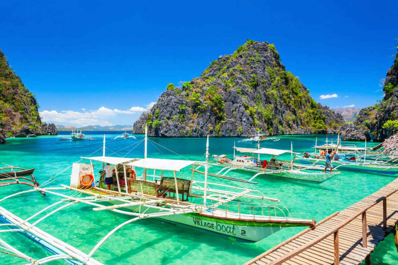 Traditional boats float in clear turquoise water near rocky islands under a bright blue sky. A wooden walkway extends from the bottom right corner.