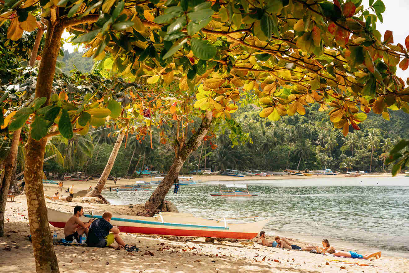 People relax on a sandy beach with boats nearby, surrounded by lush trees and calm water.