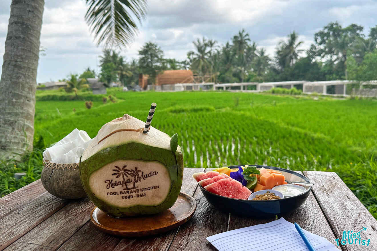 A fresh coconut with a branded logo 'Kelapa Muda Pool Barab Kitchen Ubud - Bali' on a table overlooking lush green rice paddies in Ubud, Bali