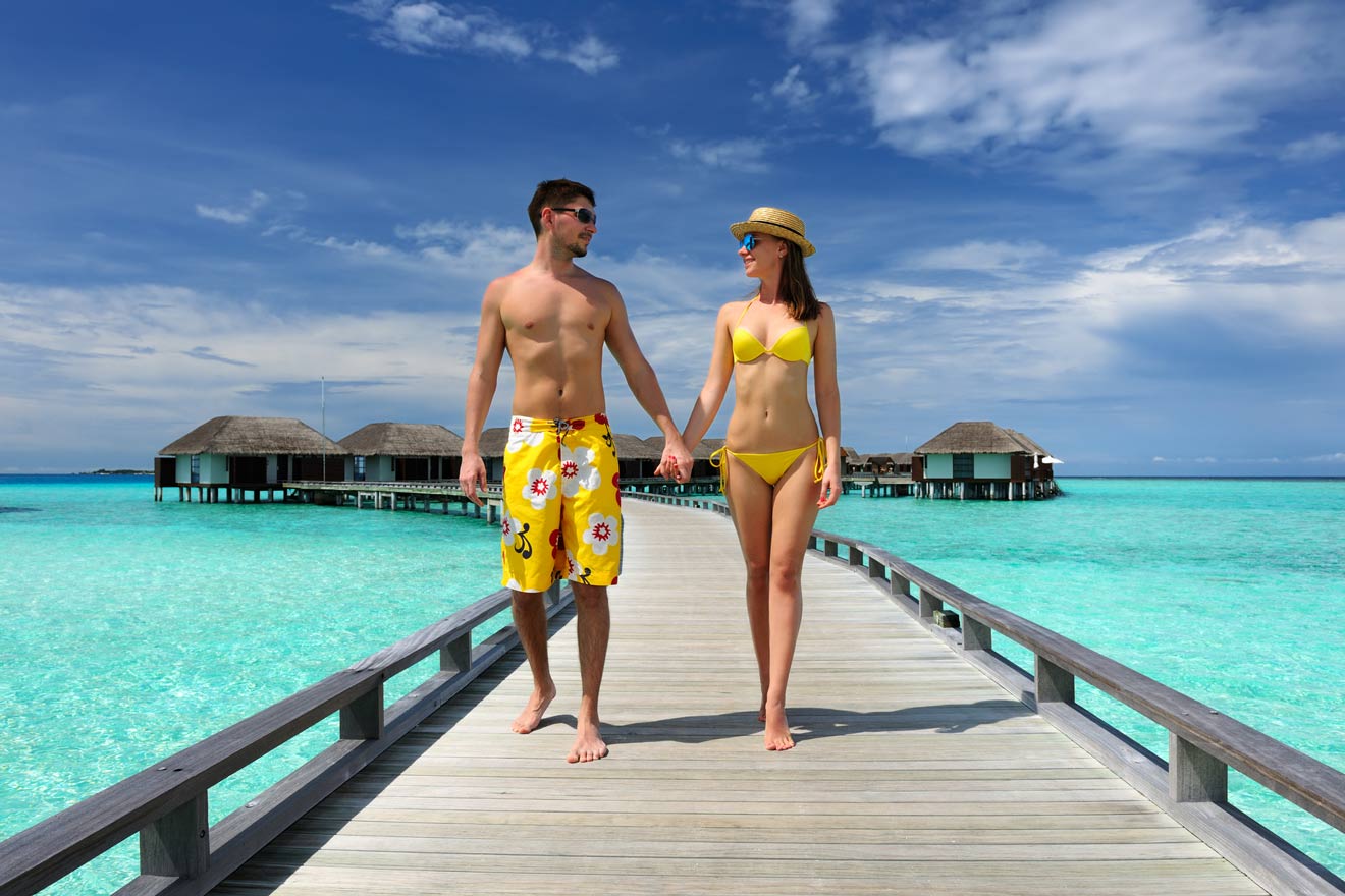 A couple in swimwear holds hands while walking on a dock over clear turquoise water, with overwater bungalows and a partly cloudy sky in the background.