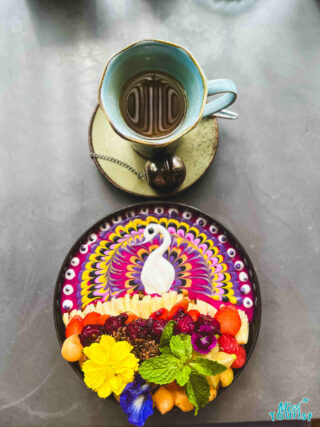 Colorful smoothie bowl garnished with edible flowers and a swan design, served next to a cup of coffee on a marble table in Ubud