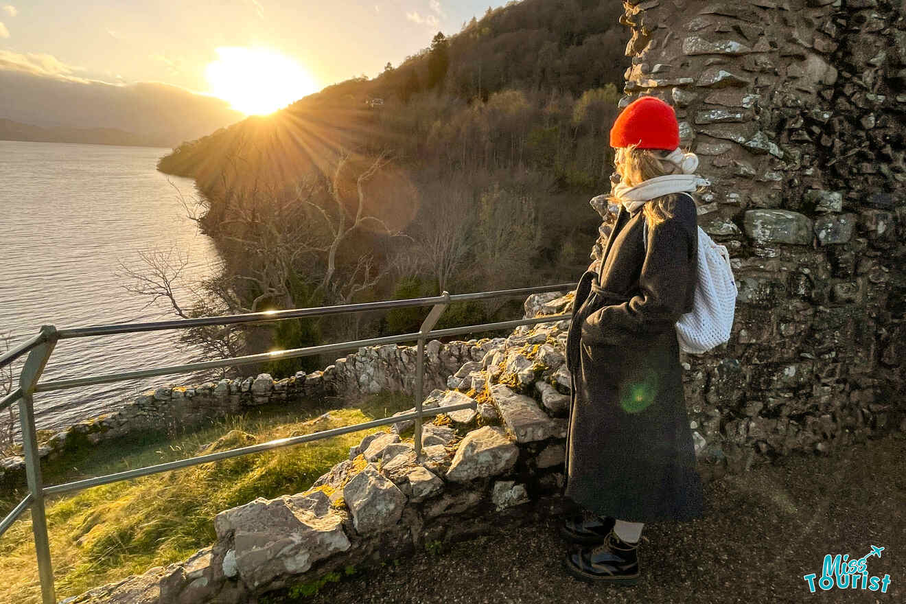  the author of the posts wearing a red beanie and dark coat looks out at a sunset over a body of water from a rocky vantage point.