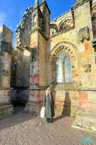 The writer of the post stands near the intricately detailed stone walls and arched windows of Rosslyn Chapel, basking in sunlight with a shadow cast on the ground