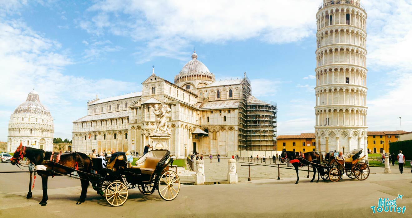 Panoramic view of Piazza dei Miracoli in Pisa, Italy, featuring the Leaning Tower of Pisa, Pisa Cathedral, and horse-drawn carriages in the foreground under a clear sky.