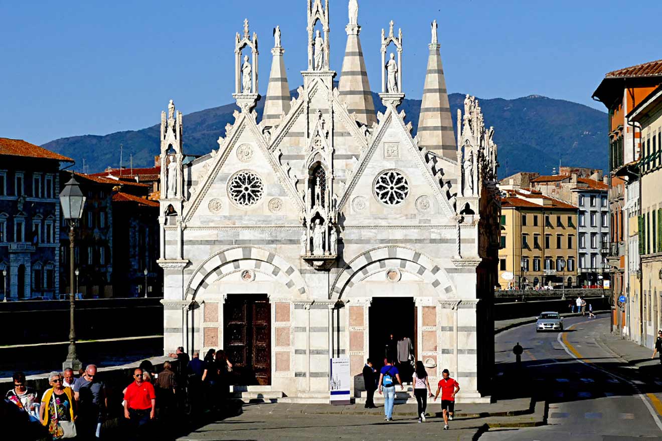 People walking by a small, ornate gothic church with pointed spires and rose windows in an urban setting with mountains in the background.