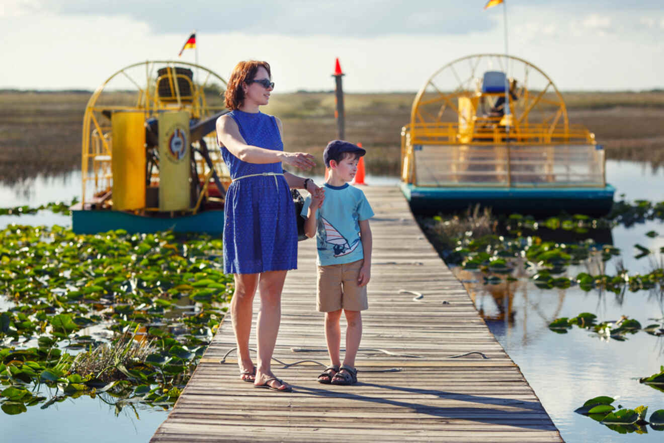 A woman and a child stand on a wooden dock surrounded by water and lily pads, with two airboats in the background in Everglades National Park 