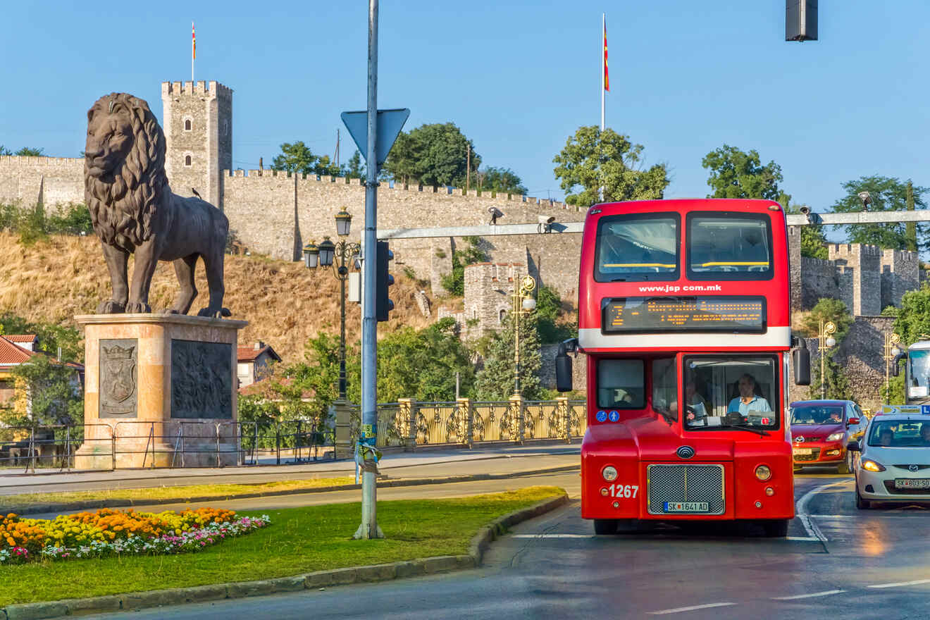 A red double-decker bus drives past a large lion statue on a city street, with a historical fortress in the background.