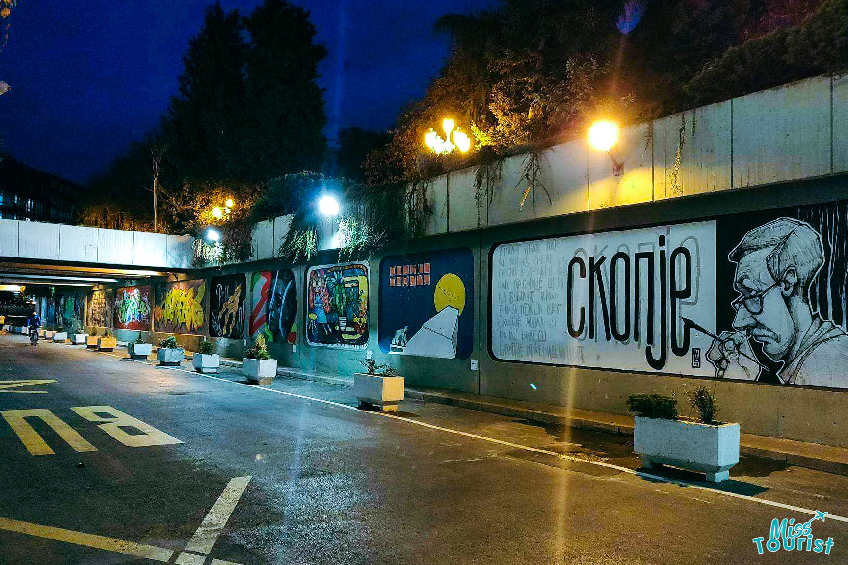 Street art on a tunnel wall at night, featuring various colorful murals, including a large "Skopje" graphic and a portrait.