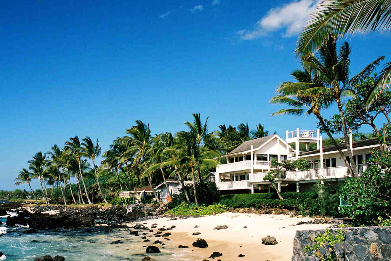 A beachfront house with palm trees, rocky shoreline, and a bright blue sky in the background.