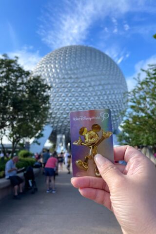 Epcot Park entrance with disney world ticket in the front and Spaceship Earth on the background and tourists 