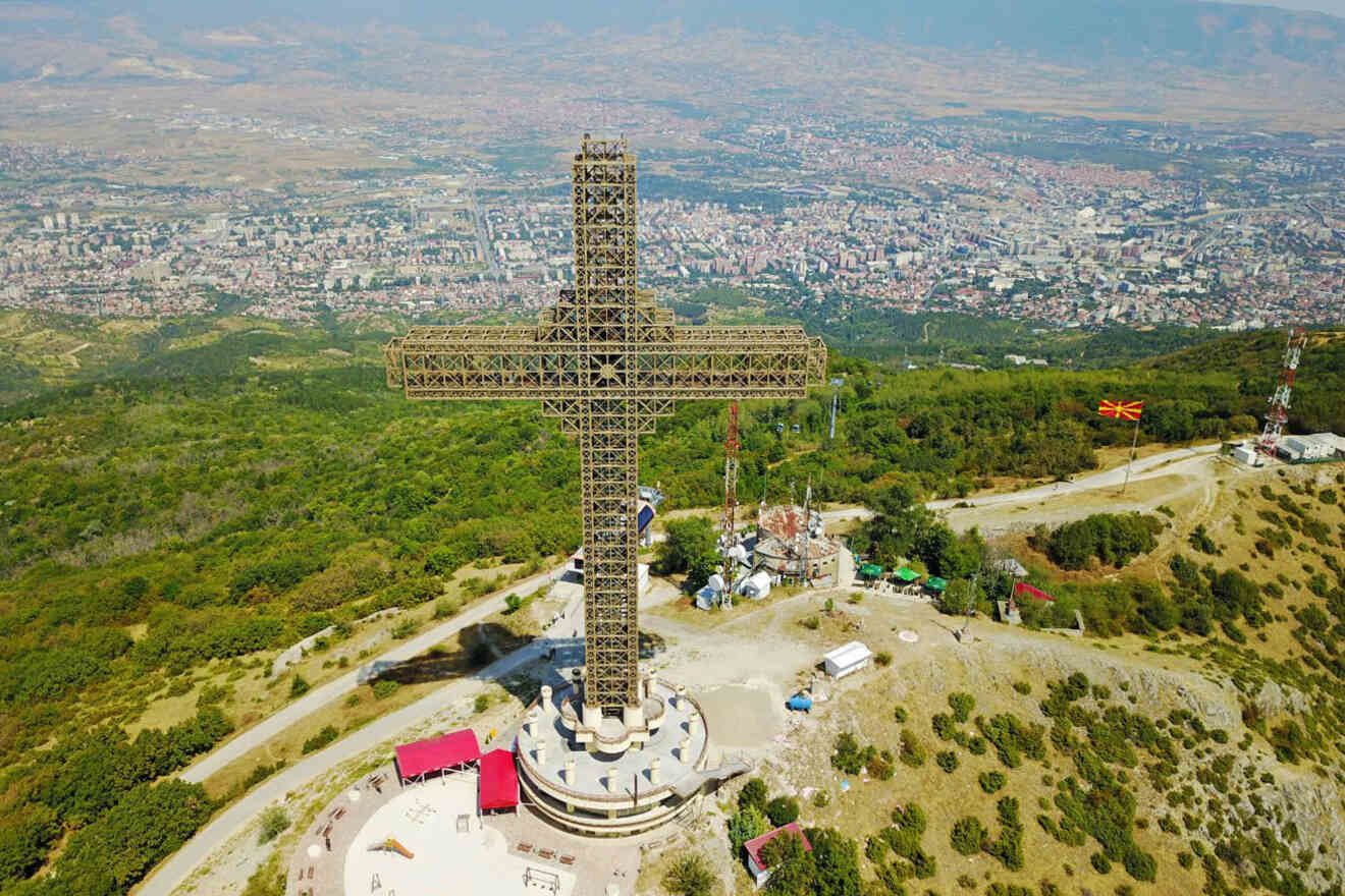 Aerial view of a large cross-shaped structure on a hilltop overlooking a city, surrounded by lush green landscape and roads, with a few smaller buildings nearby.