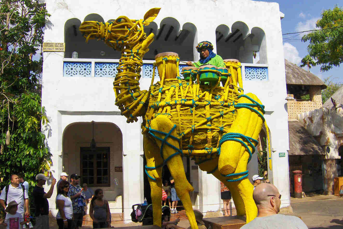 A person sits atop a large yellow camel sculpture made of rope, surrounded by people in an outdoor area with white buildings.