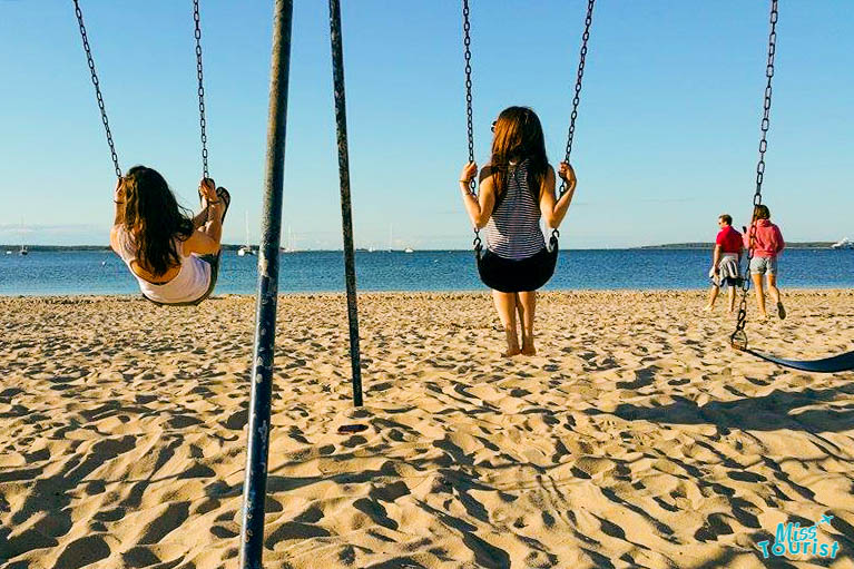 Two people enjoying swings on Coopers Beach in Hampton, overlooking the calm ocean, with footprints in the sand and another couple strolling in the background on a clear day.