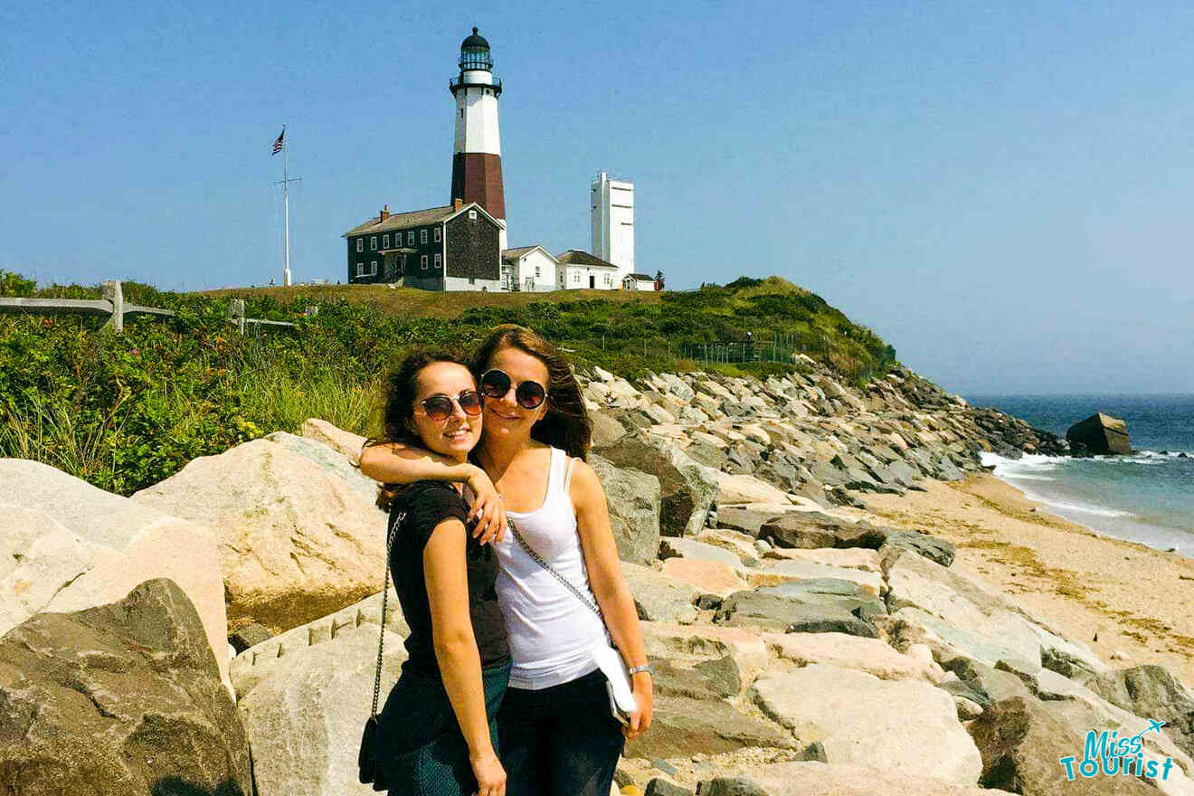 Two smiling women in sunglasses posing in front of the historic Montauk Point Lighthouse, with rocky beach foreground and clear blue skies, captured on a sunny day.