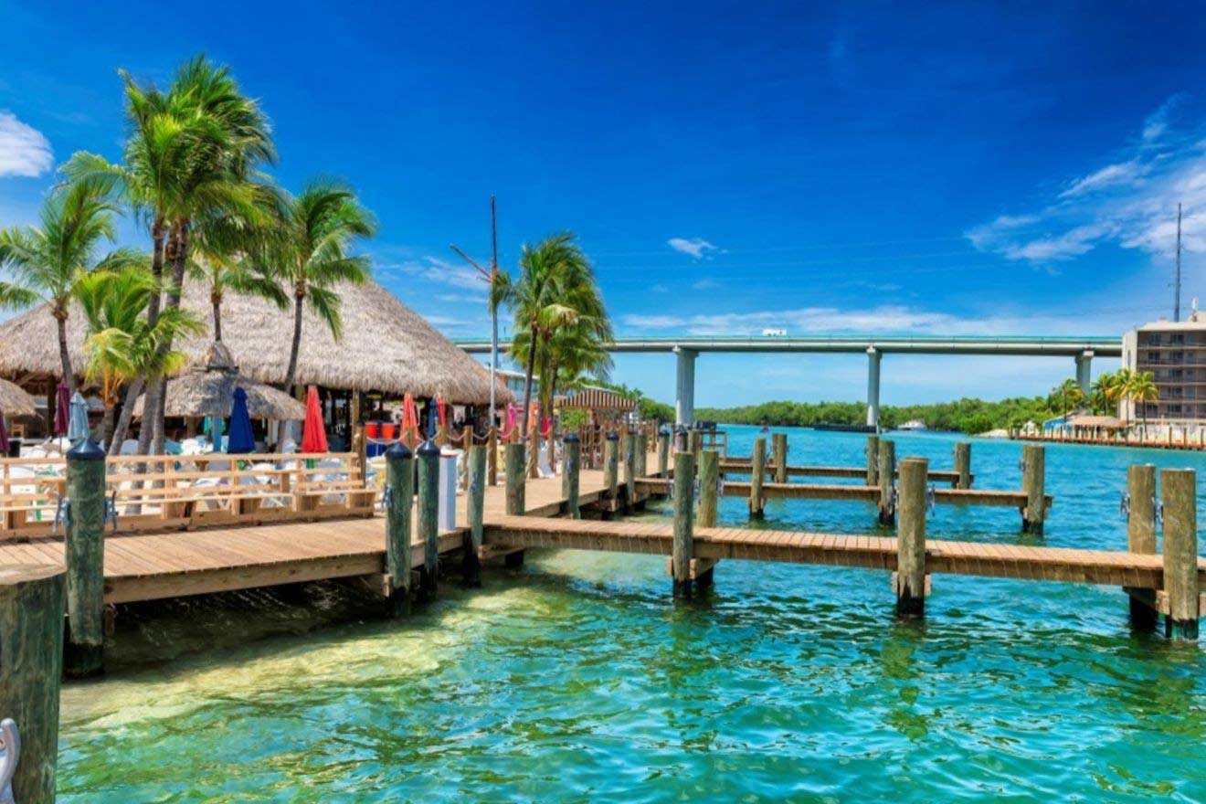 A wooden pier extends over a clear, turquoise body of water, leading to a thatched-roof structure and seating area with umbrellas. A bridge and lush greenery are visible in the background under a bright blue sky.