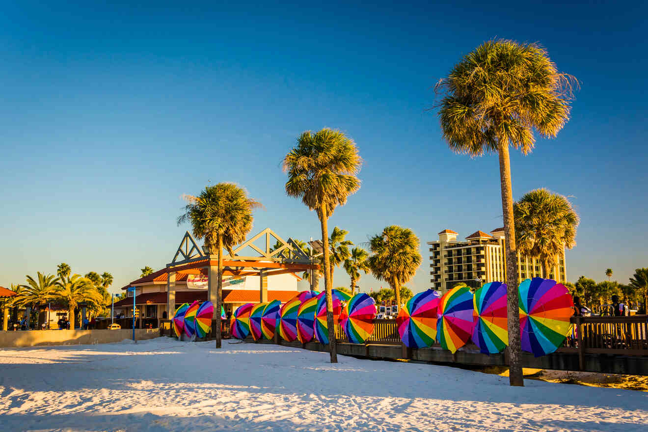 Colorful beach umbrellas line a sandy beach with palm trees and a building in the background under a clear blue sky.